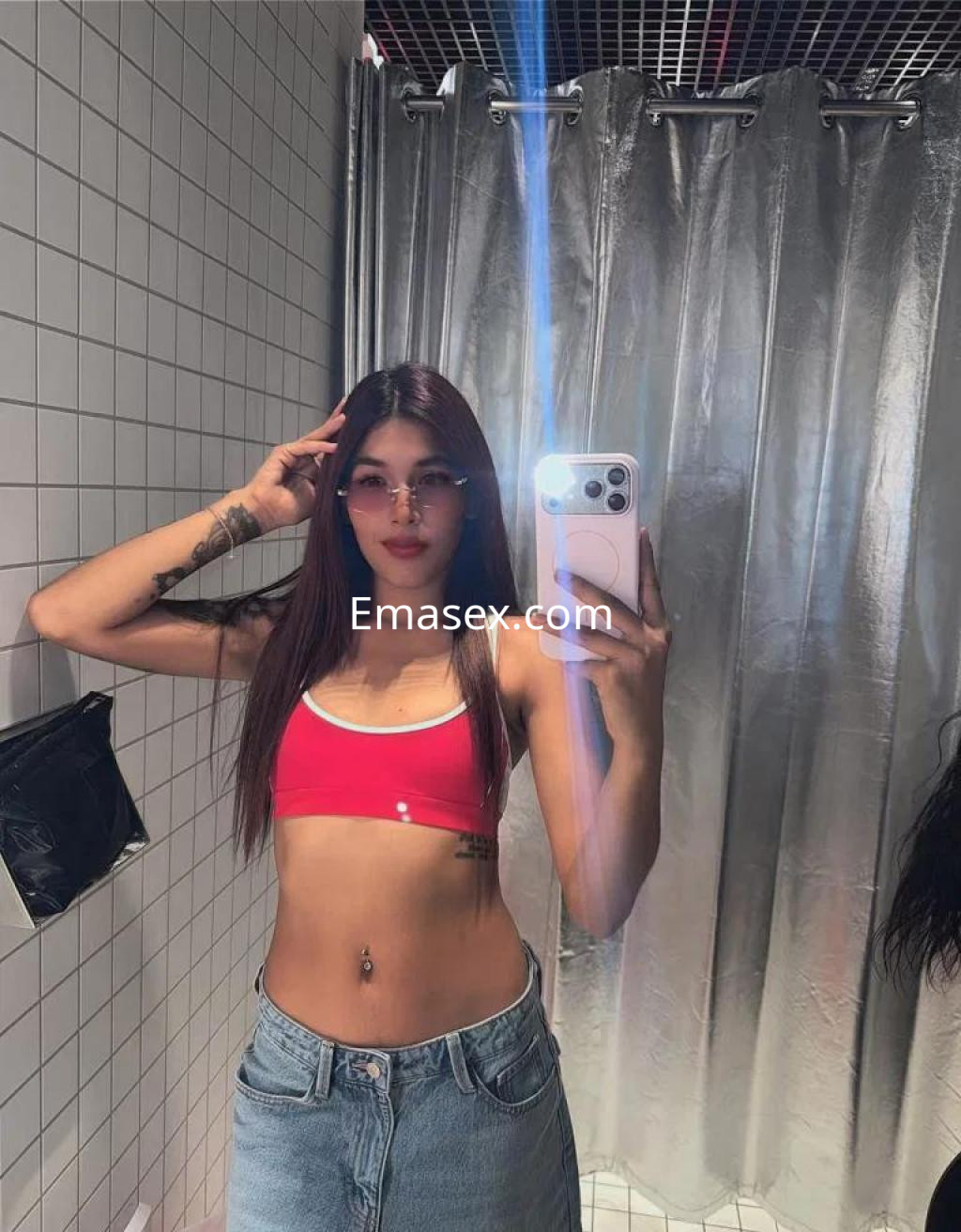 Escort en Girona con top rojo y pantalones de mezclilla, posando frente a un espejo en un ambiente moderno.
