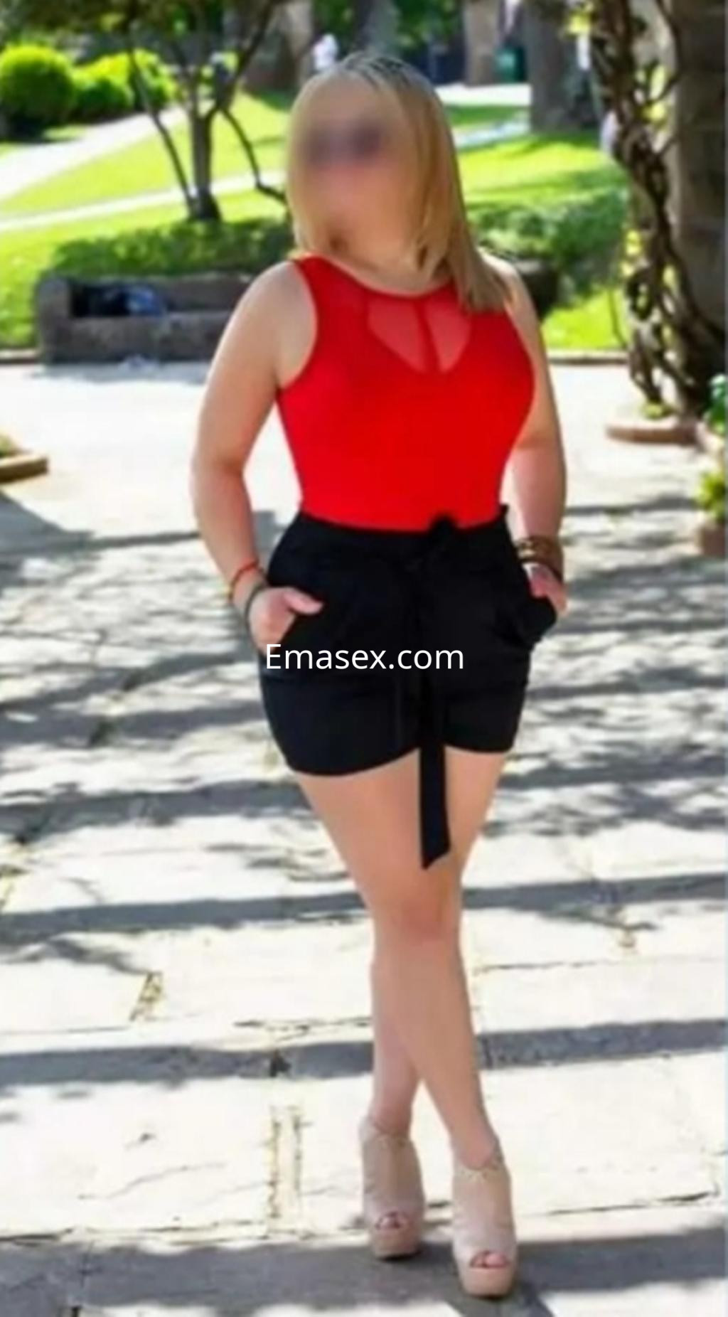 Escort en Santander viste un body rojo con detalles en el escote y pantalones cortos negros, posando en un parque.