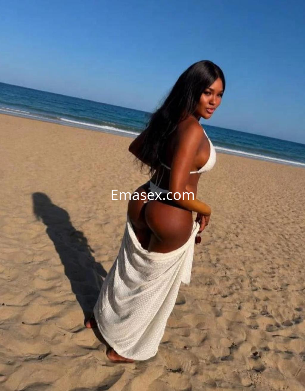 La escort en Madrid se muestra en la playa, vistiendo un bikini blanco y una falda larga, con el mar de fondo.