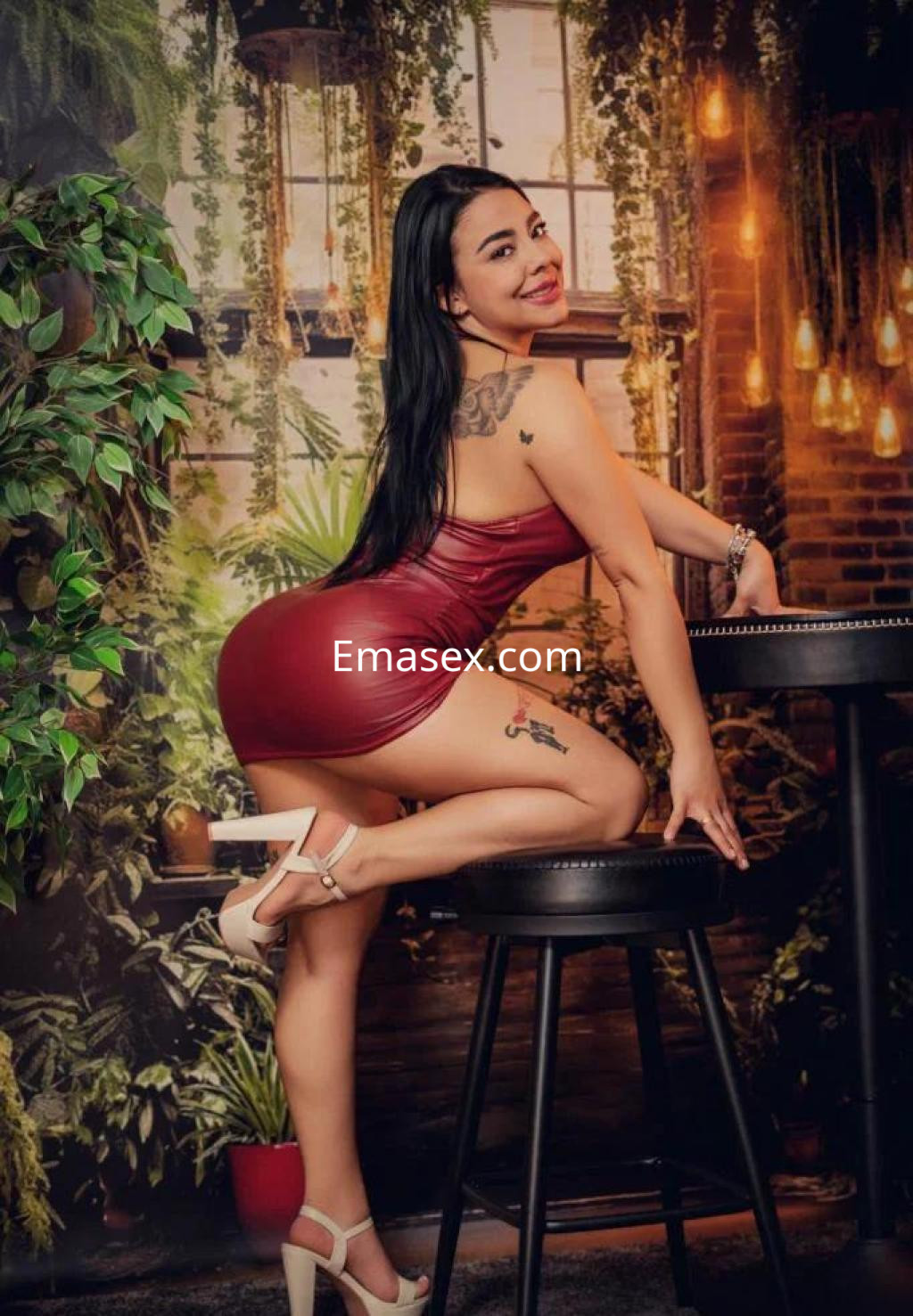 Escort posando en un vestido rojo ajustado y tacones, con fondo de plantas en Madrid.