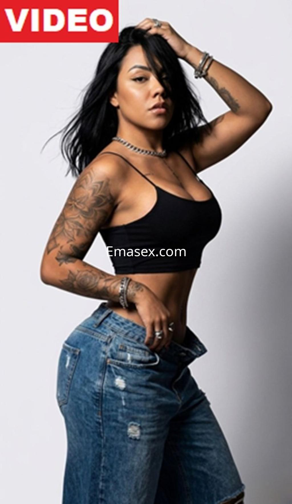 La escort en Pamplona luce una camiseta negra sin mangas y jeans rotos, mostrando sus tatuajes y una pose confiada.
