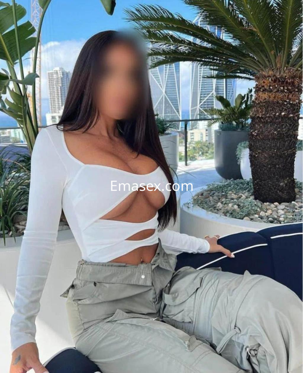 Escort en Tenerife con blusa blanca de corte asimétrico y pantalones claros, sentada en un entorno moderno.