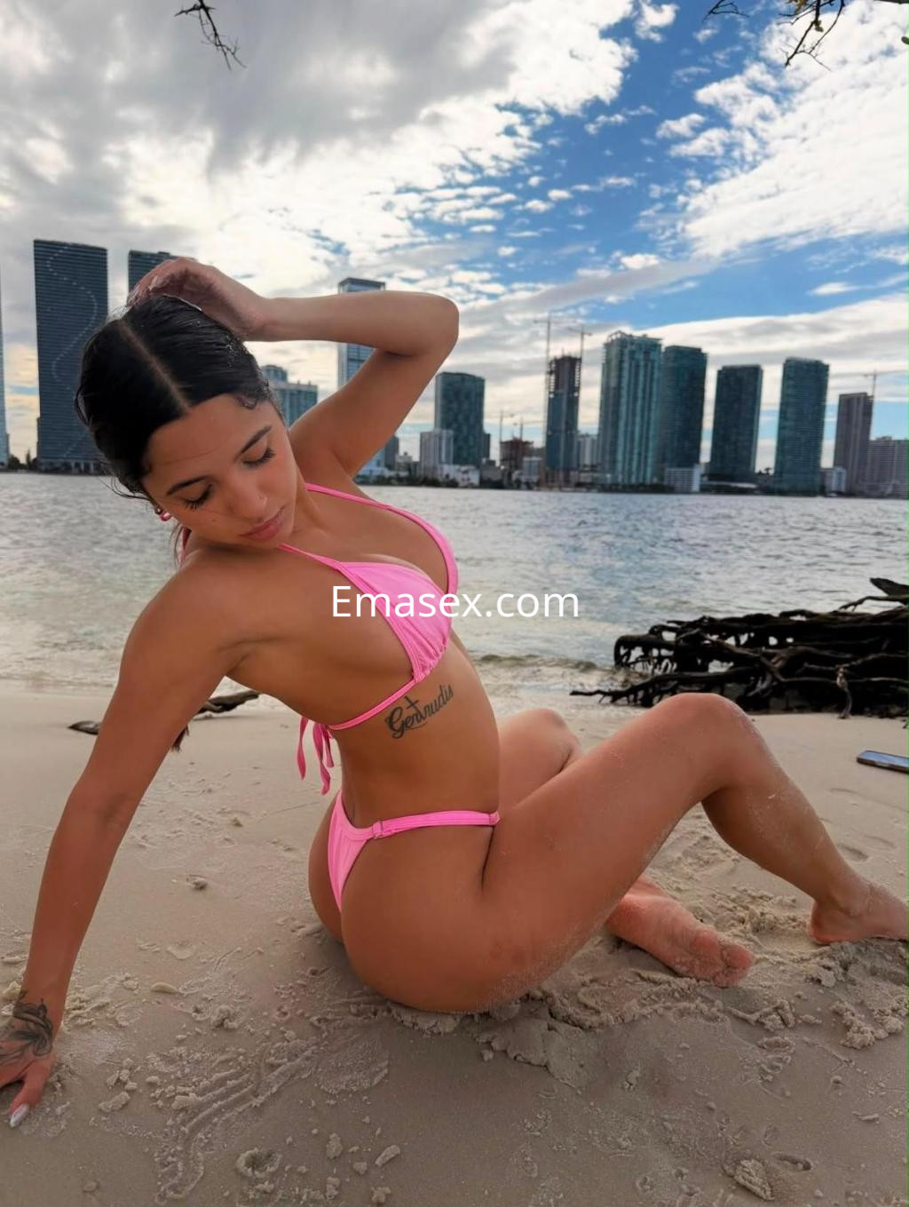 Escort en Donosti luciendo un bikini rosa brillante, sentada en la arena junto a un paisaje urbano moderno.