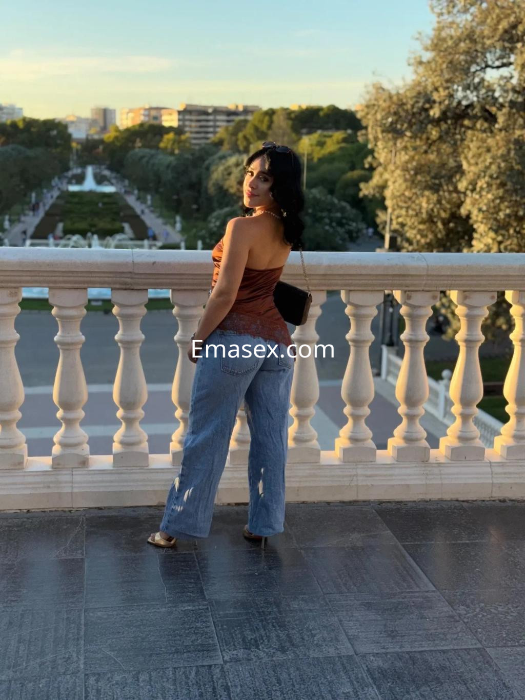 Puta posando de espaldas en un balcón, lleva una blusa marrón sin mangas y jeans claros, con paisaje urbano al fondo en Zaragoza.