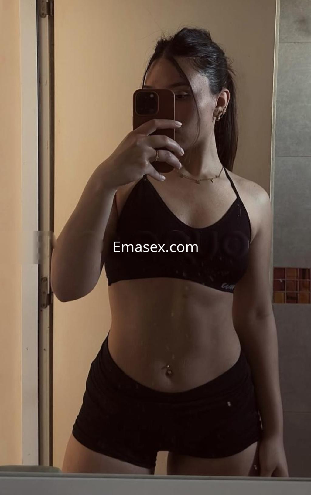 Escort en Barcelona se toma un selfie en el espejo, vistiendo un sujetador negro y shorts cortos oscuros.