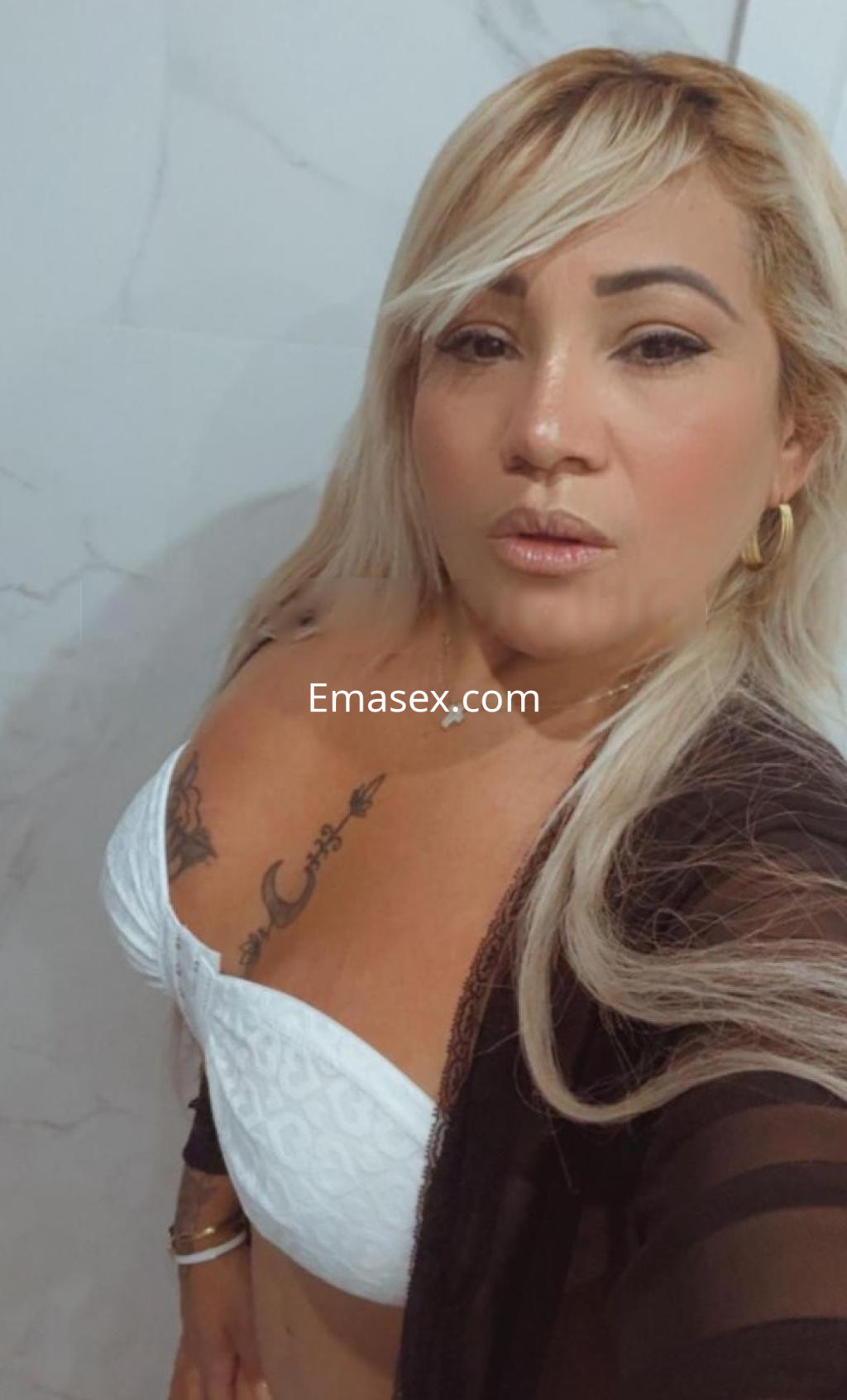 Escort en Barcelona posa en un baño, vistiendo un sujetador blanco y una bata negra transparente, con cabello largo y rubio.