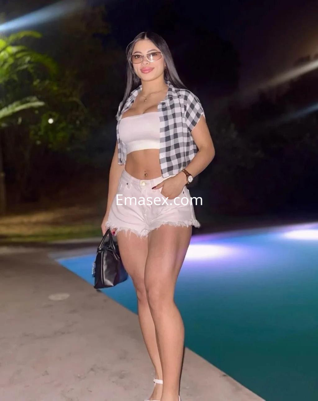 La puta está de pie junto a una piscina en Burgos, vistiendo un top blanco y shorts cortos, con gafas y cabello largo.