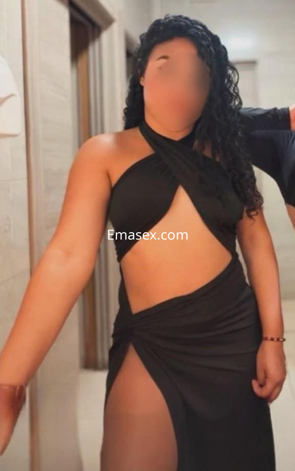 Escort en Barcelona con cabello rizado, viste un vestido negro con cortes laterales, posando en un ambiente interior.