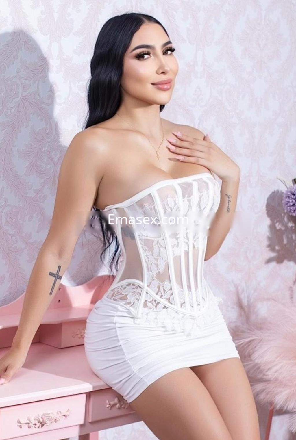 Puta en Barcelona vistiendo un corset blanco con encaje y una falda ajustada, posando con una mano en su pecho.