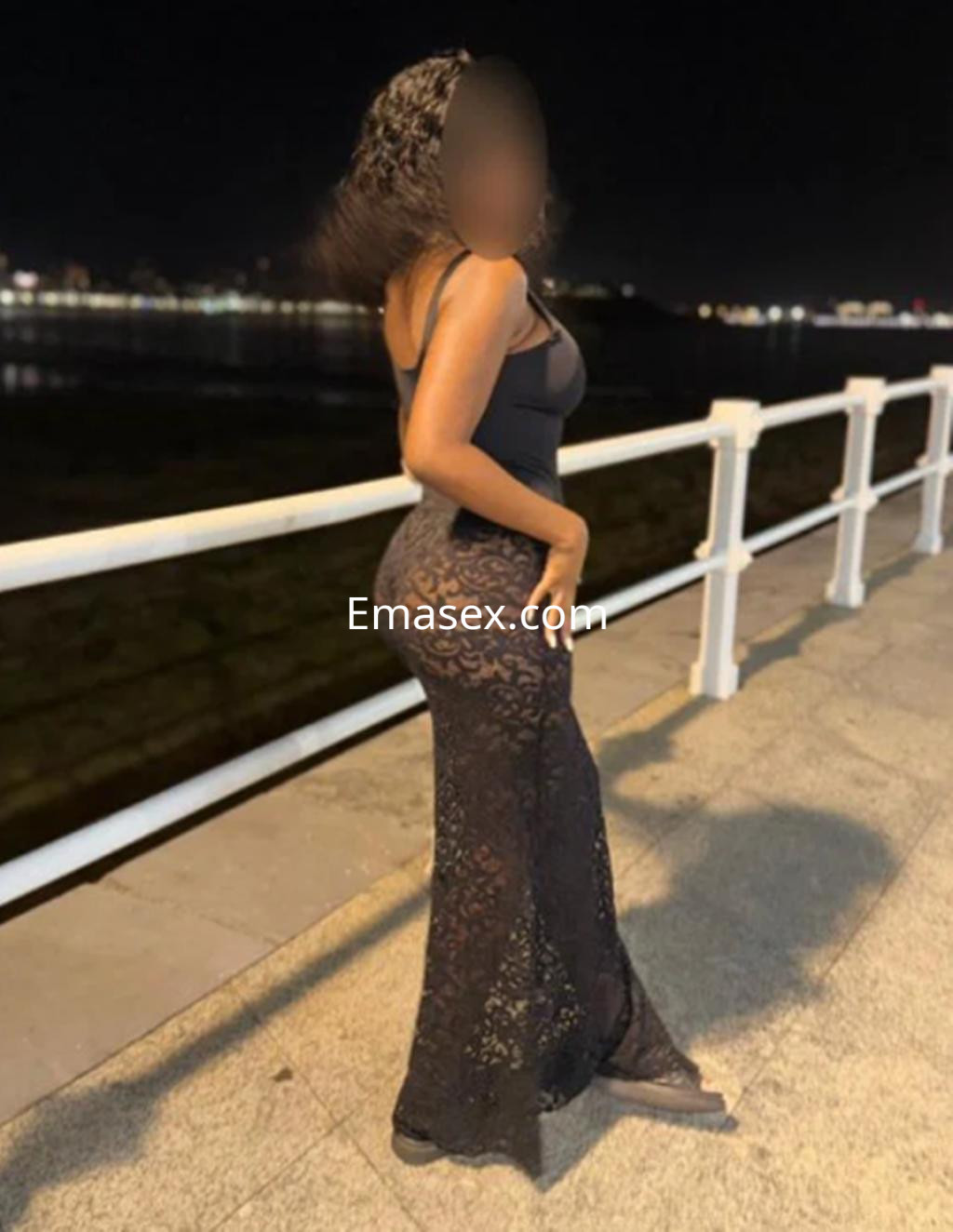 Puta en Gijón, de pie en un paseo marítimo nocturno, lleva un top negro y una falda larga de encaje negra.