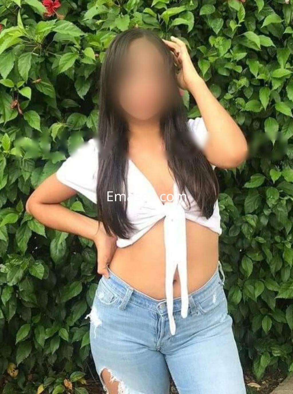 Escort en Barcelona con blusa blanca atada y jeans desgastados, posando con una mano en la cadera entre vegetación.