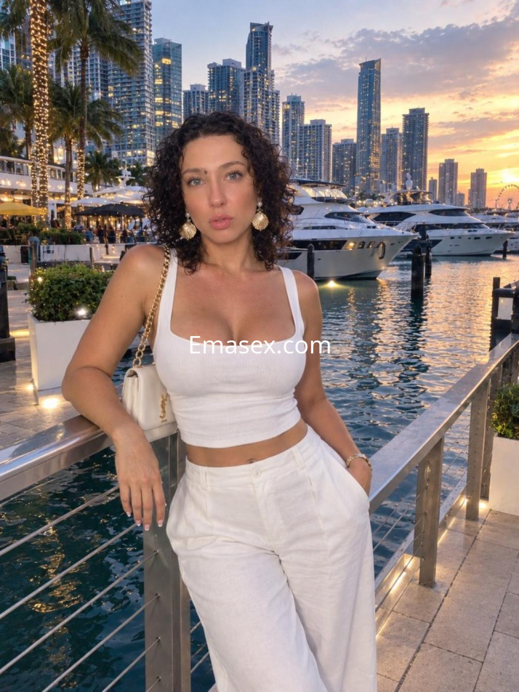Escort trans en Logroño con cabello rizado y oscuro, viste un top blanco y pantalones claros, posando junto al agua.