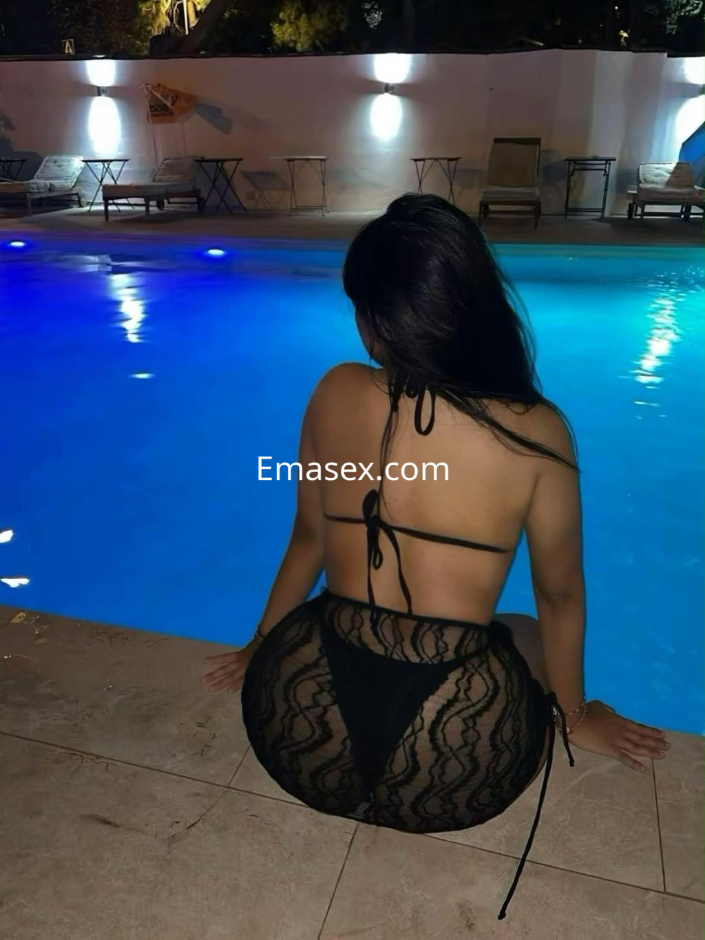 Puta de espaldas junto a una piscina en Santander, con lencería negra y cabello largo, posando con confianza.