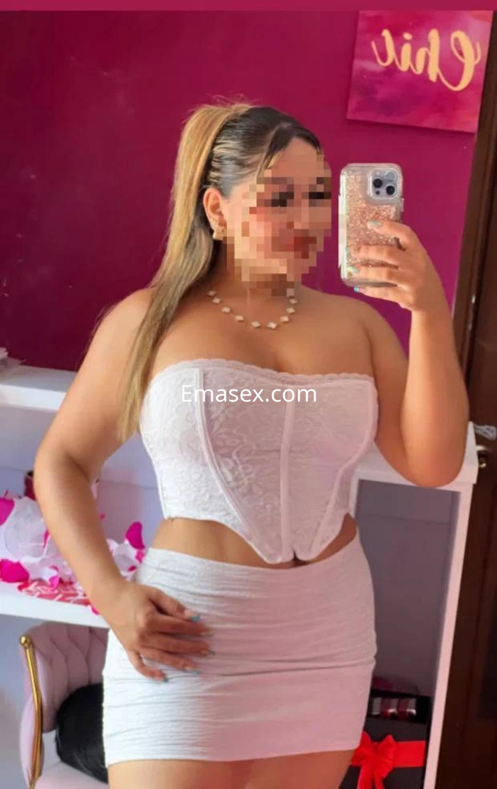 Puta en Murcia posando frente al espejo con un corset blanco y una minifalda ajustada, luciendo un peinado recogido.