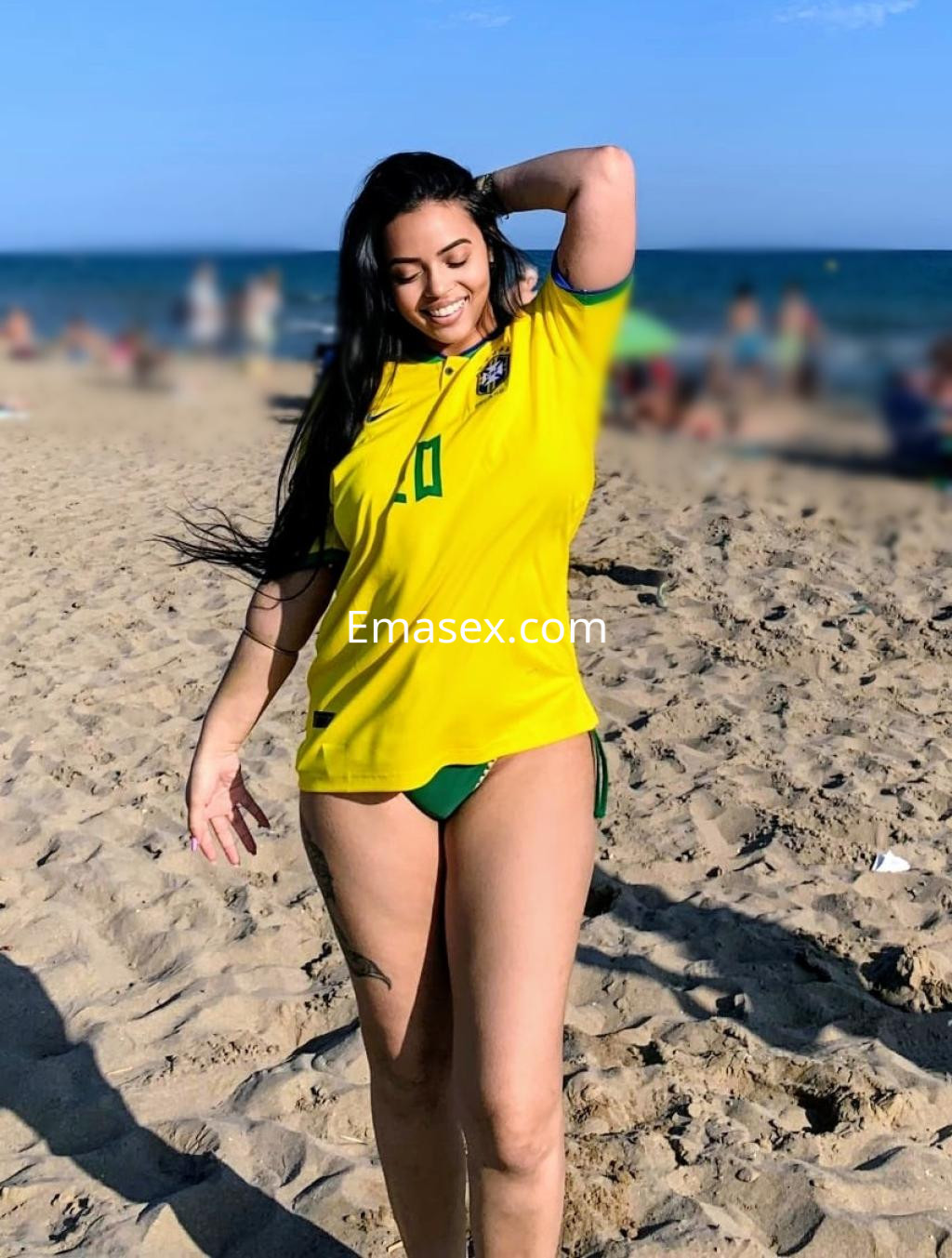 Puta en Pamplona con cabello largo y oscuro, luciendo una camiseta amarilla y bikini verde en la playa.