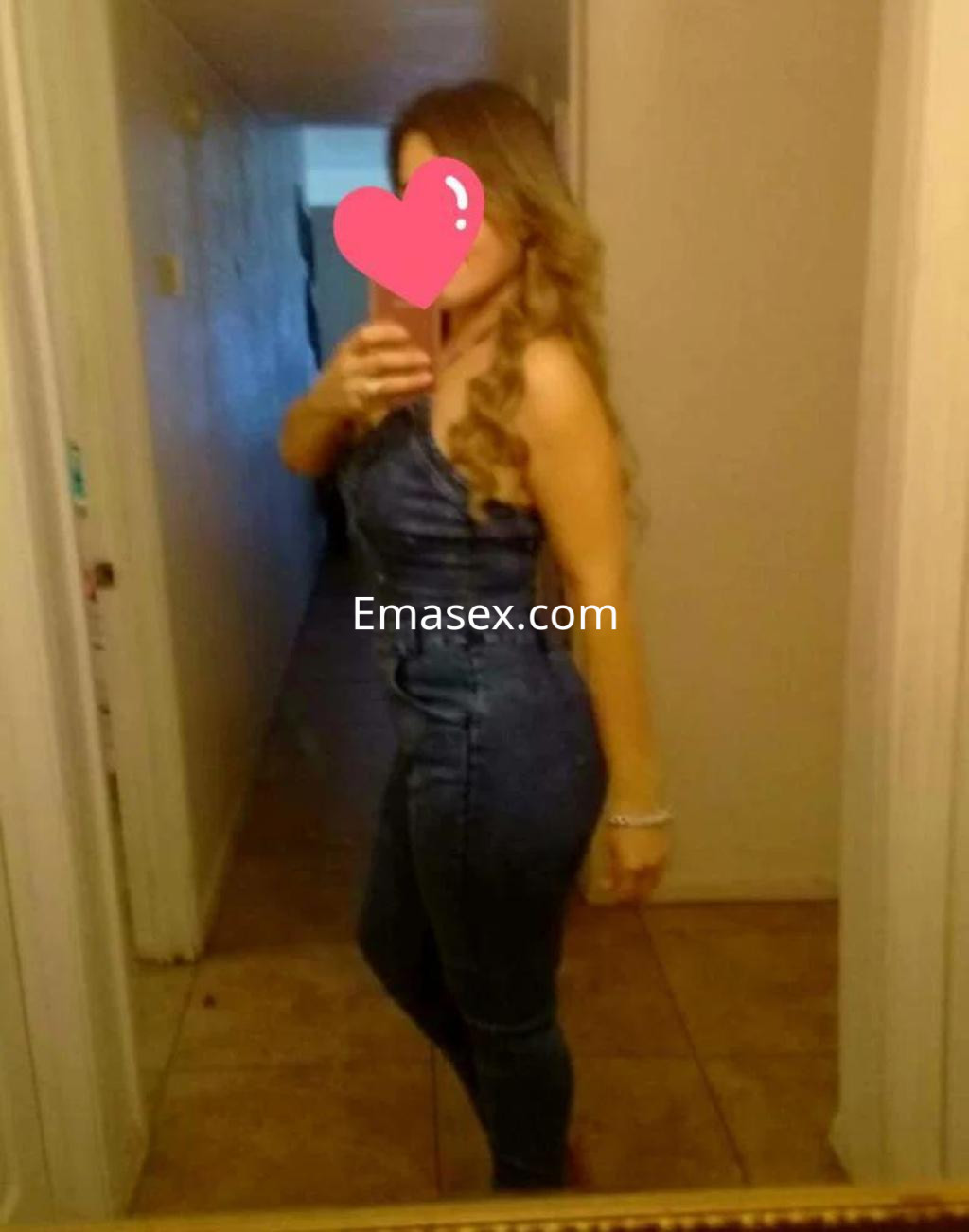 Puta en Salamanca se toma una selfie en un espejo, luciendo un ajustado overol de mezclilla y cabello ondulado.