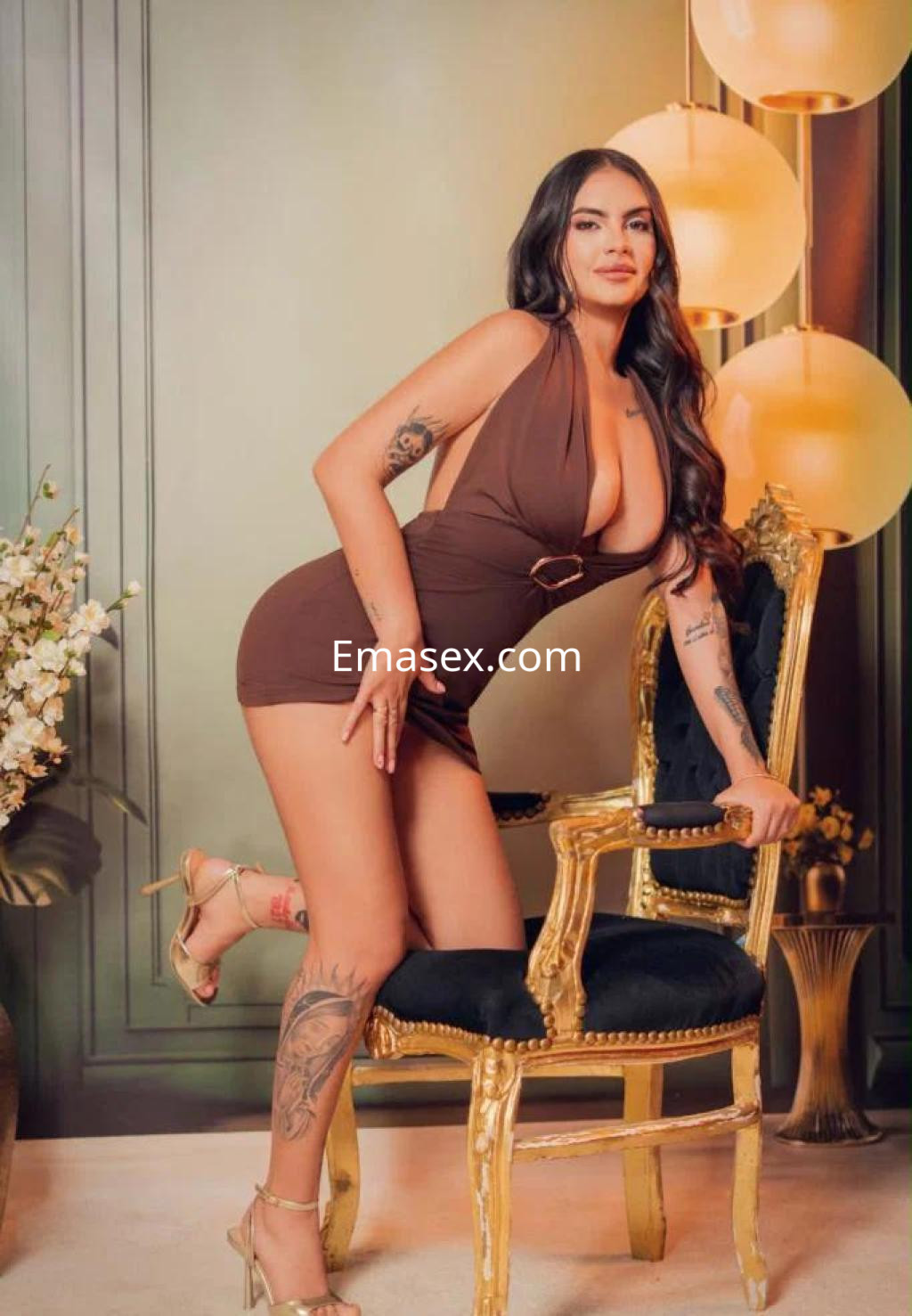 Escort posando de pie junto a una silla dorada, vistiendo un vestido marrón y tacones, en un ambiente elegante en Palma de Mallorca.