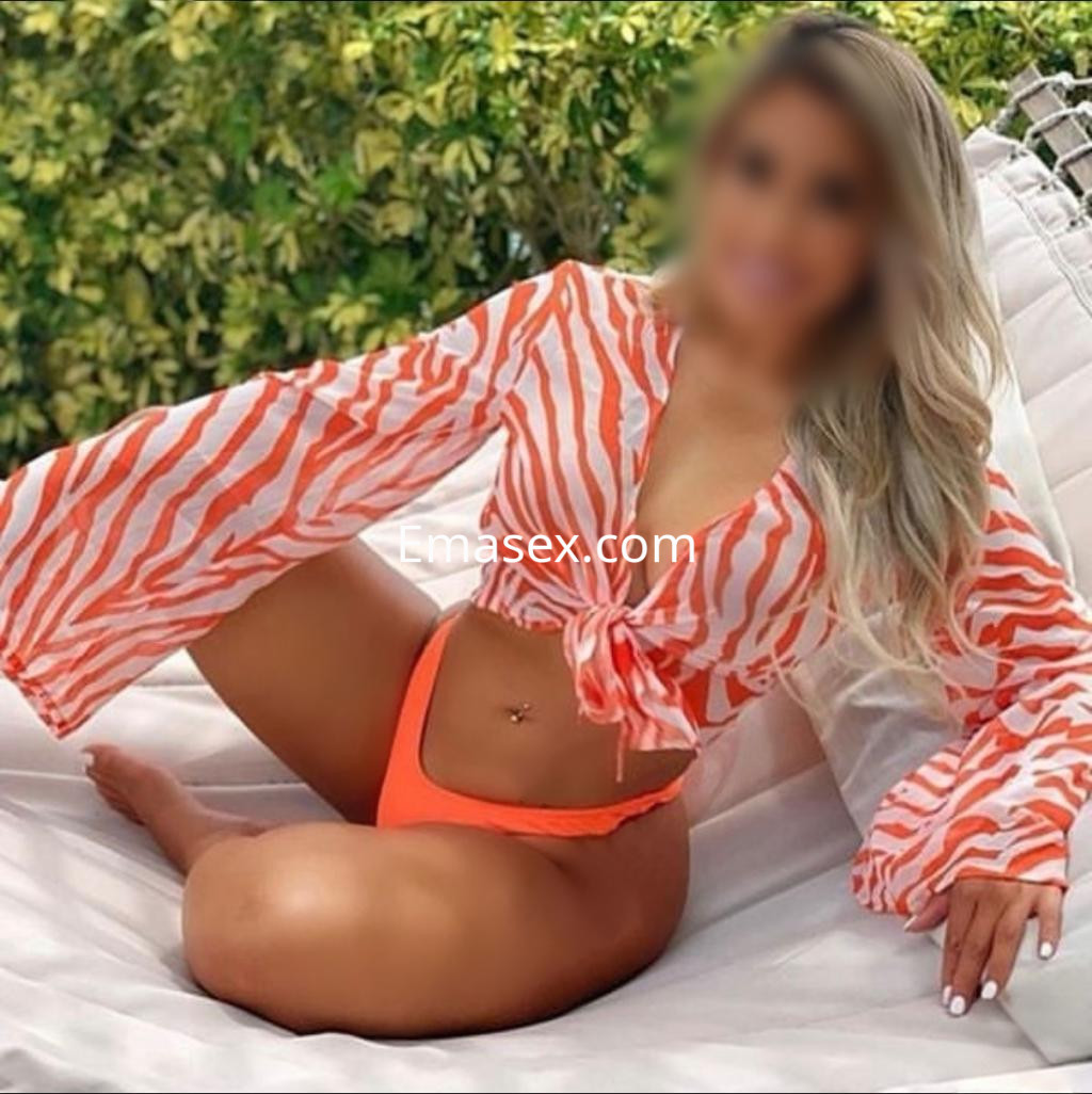 Escort en Granada con blusa de rayas naranjas y blancas, luciendo una bikini de color naranja, sentada en un entorno natural.