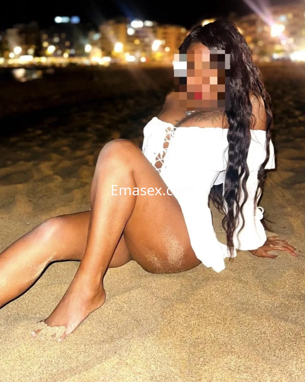 Escort en Zaragoza sentada en la arena, luciendo una blusa blanca con detalles y mostrando su piel bronceada.