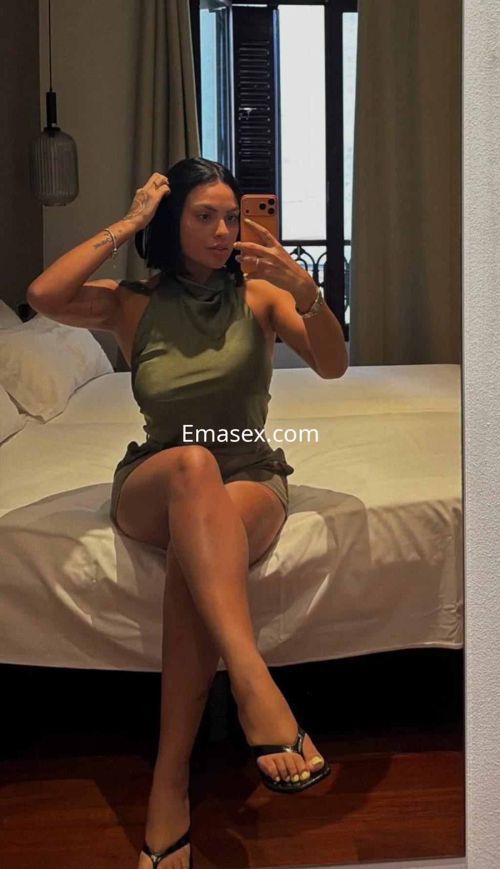 Escort en Murcia, sentada en una cama, vistiendo un vestido verde claro y sandalias, mientras toma una selfie en el espejo.