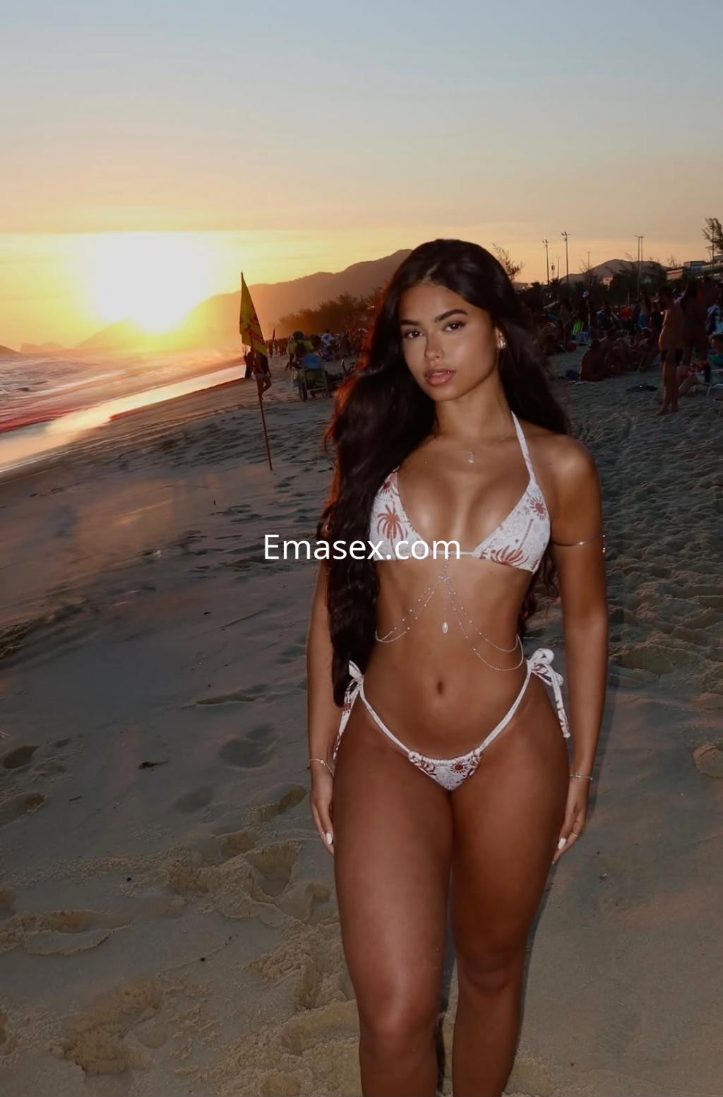 Puta en Málaga, vistiendo un bikini blanco con detalles, posando en la playa al atardecer con cabello largo y suelto.