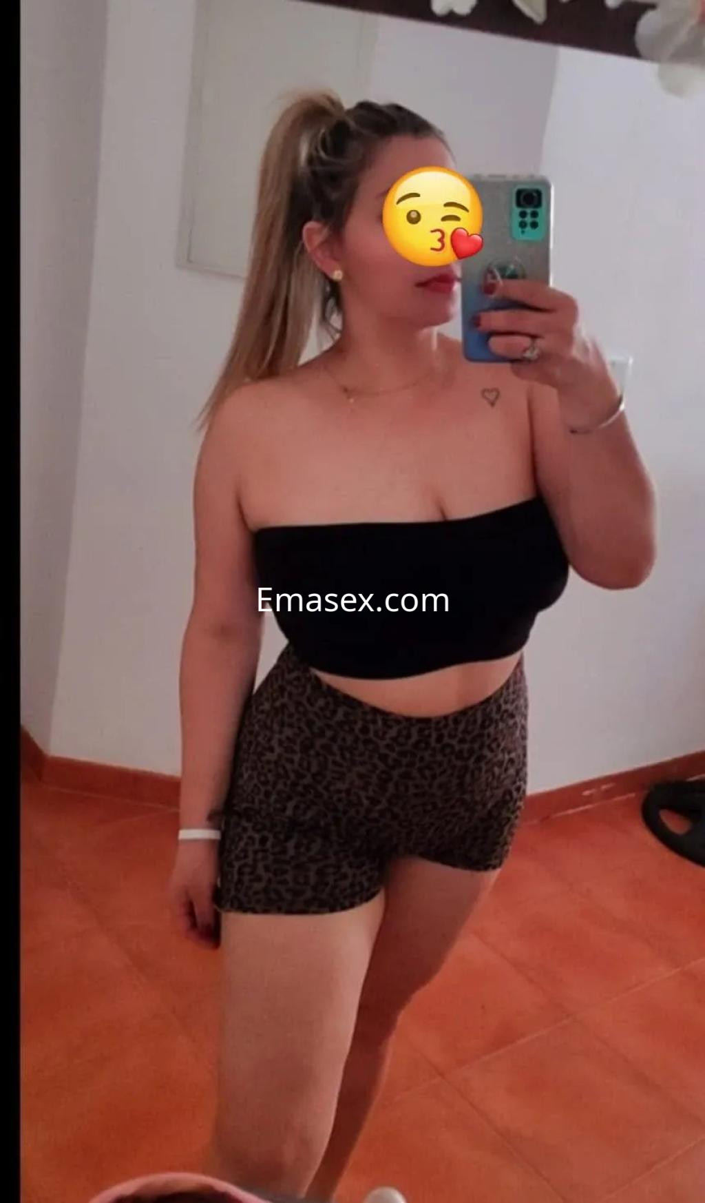 Escort en Toledo con cabello rubio recogido, lleva un top negro y shorts de leopardo, posando frente al espejo.