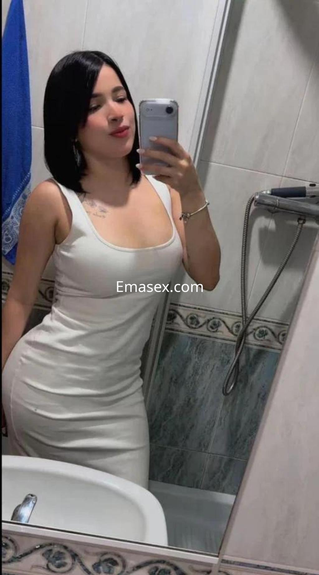 Escort en Badajoz posando frente al espejo, luciendo un vestido ajustado blanco y cabello corto oscuro.
