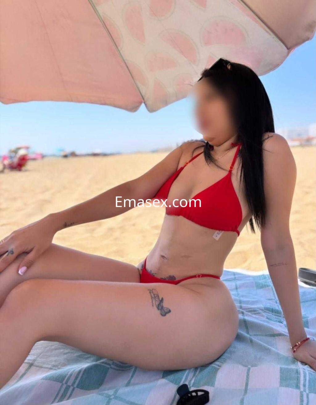 Escort sentada en la playa con un bikini rojo, bajo una sombrilla, mostrando su tatuaje y cabello largo.