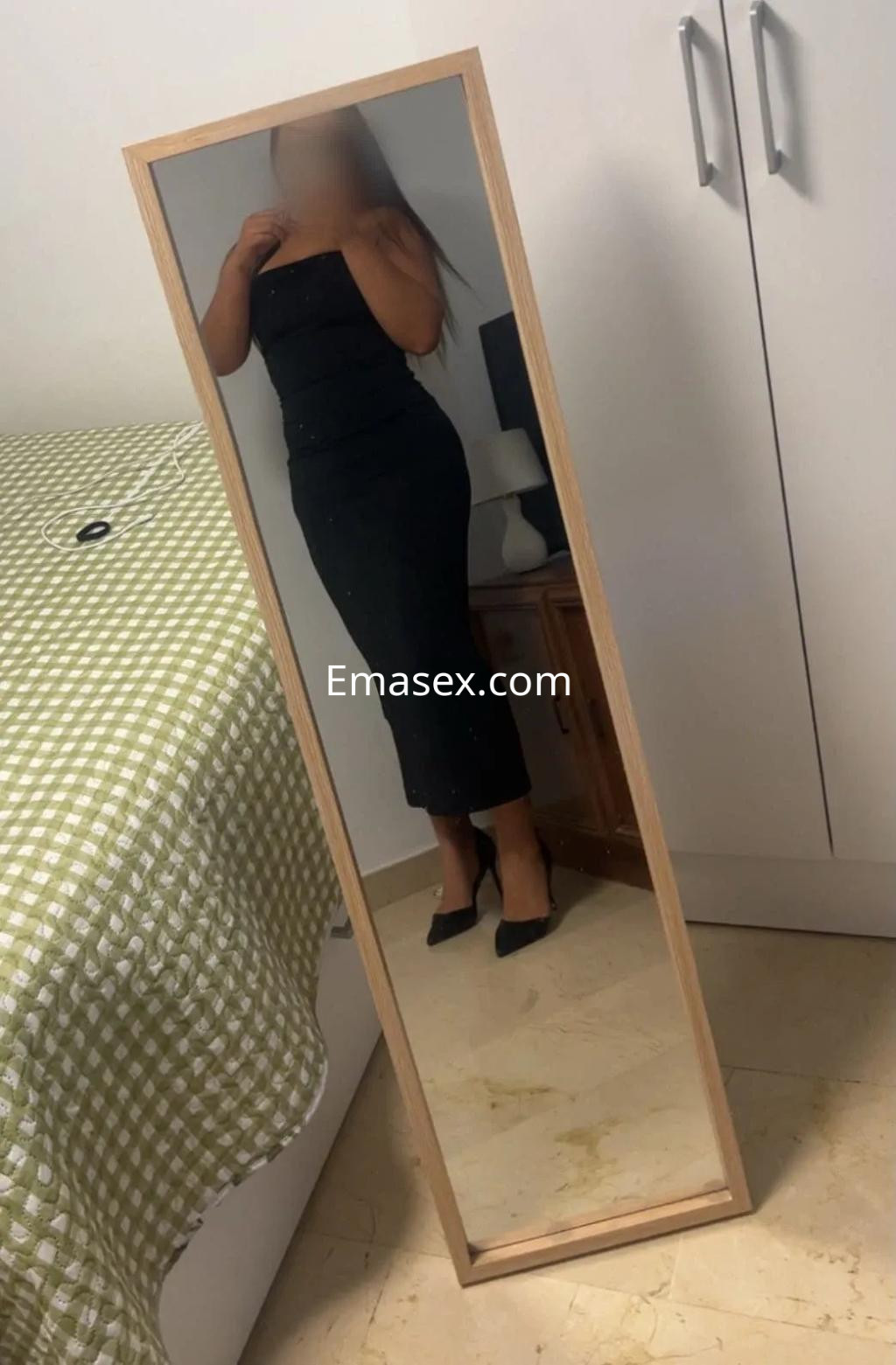 La puta en Granada viste un elegante vestido negro ajustado y tacones, posando frente a un espejo.