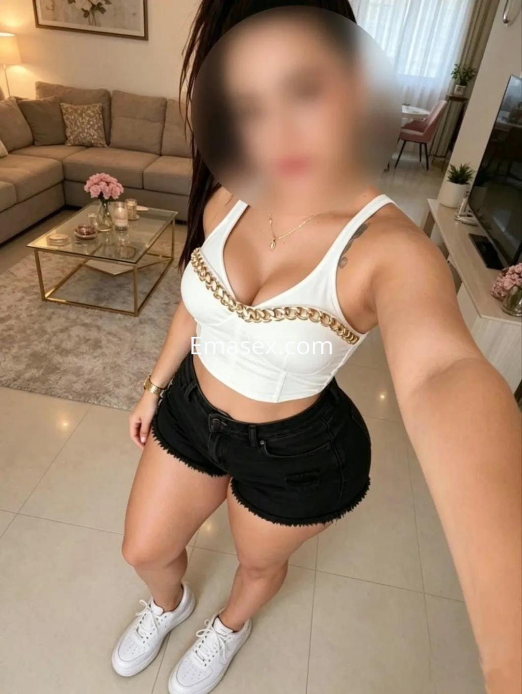 Escort en Vigo luciendo un top blanco con cadena y pantalones cortos negros en un ambiente interior moderno.