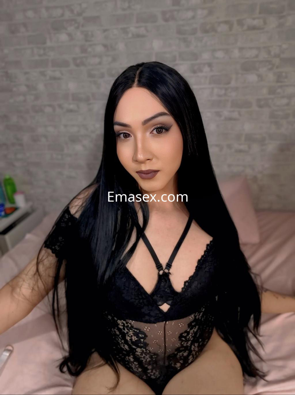 Puta trans en Pamplona, con cabello largo y lacio, viste un body negro de encaje mientras posa sentada en la cama.