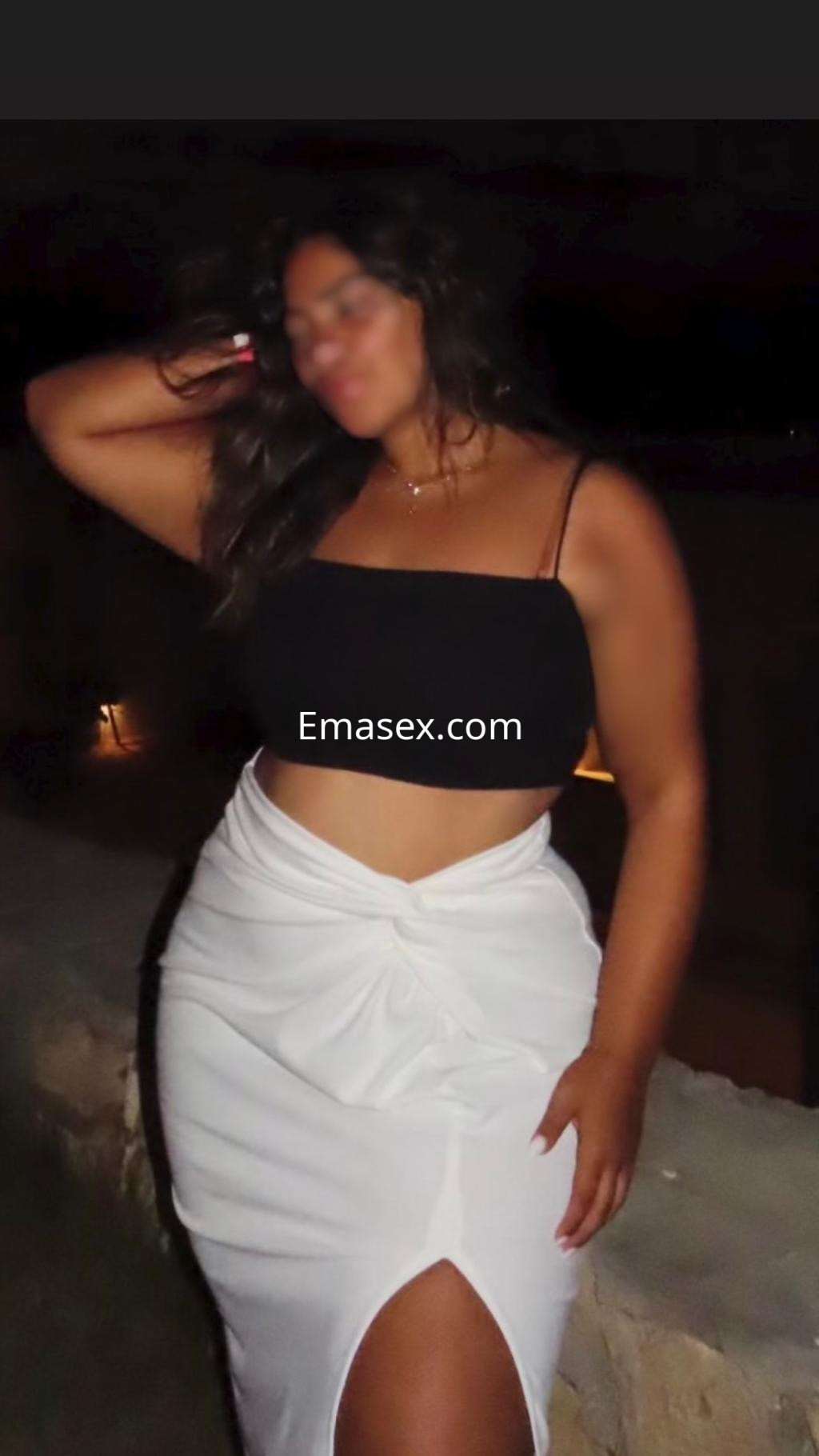 Puta posando en León, con blusa negra y falda blanca, mostrando una actitud relajada en un ambiente nocturno.