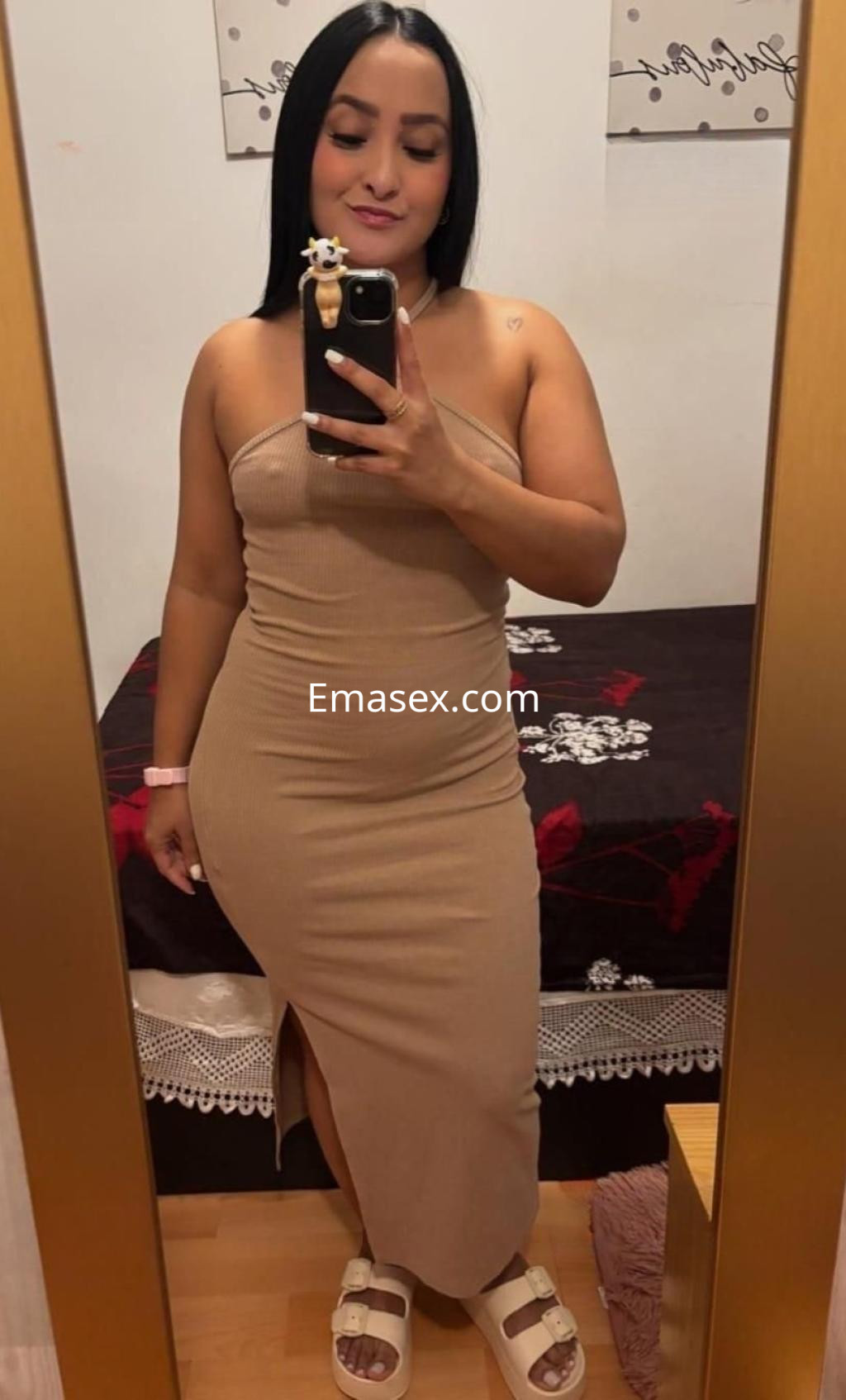 Escort en Madrid, posando frente a un espejo con un vestido ajustado color beige y sandalias blancas.