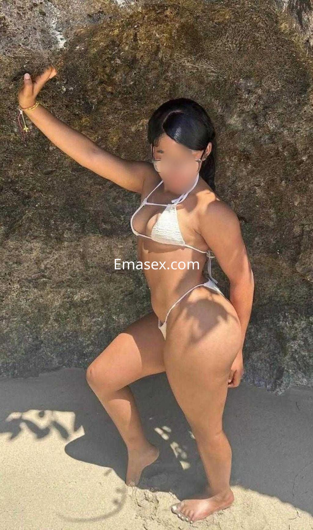 Escort en Madrid posando en la playa con un bikini blanco, apoyada en una roca, luciendo bronceado.