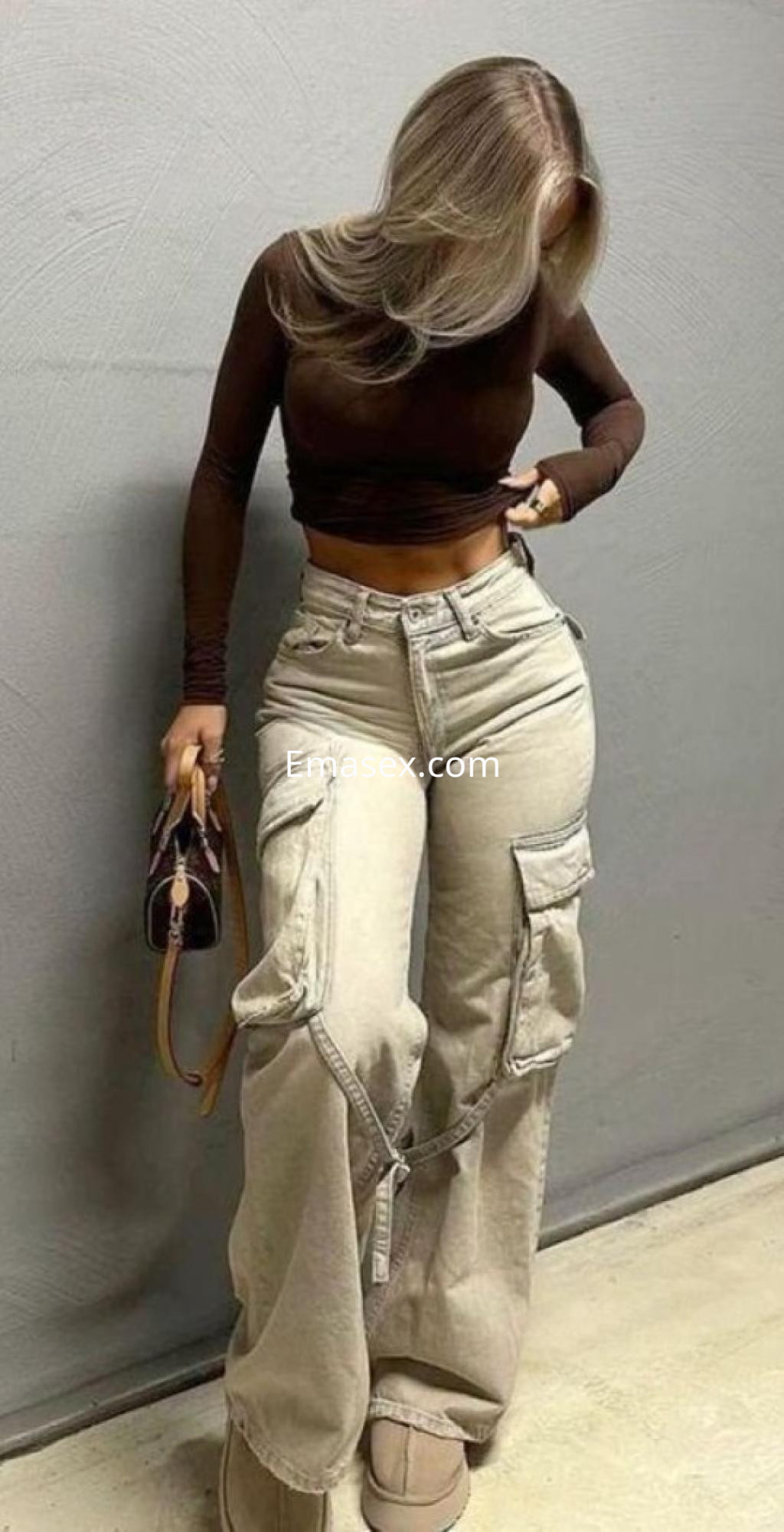 Escort con cabello rubio lacio, vistiendo un top marrón y pantalones cargo claros, ajustándose la ropa en Madrid.