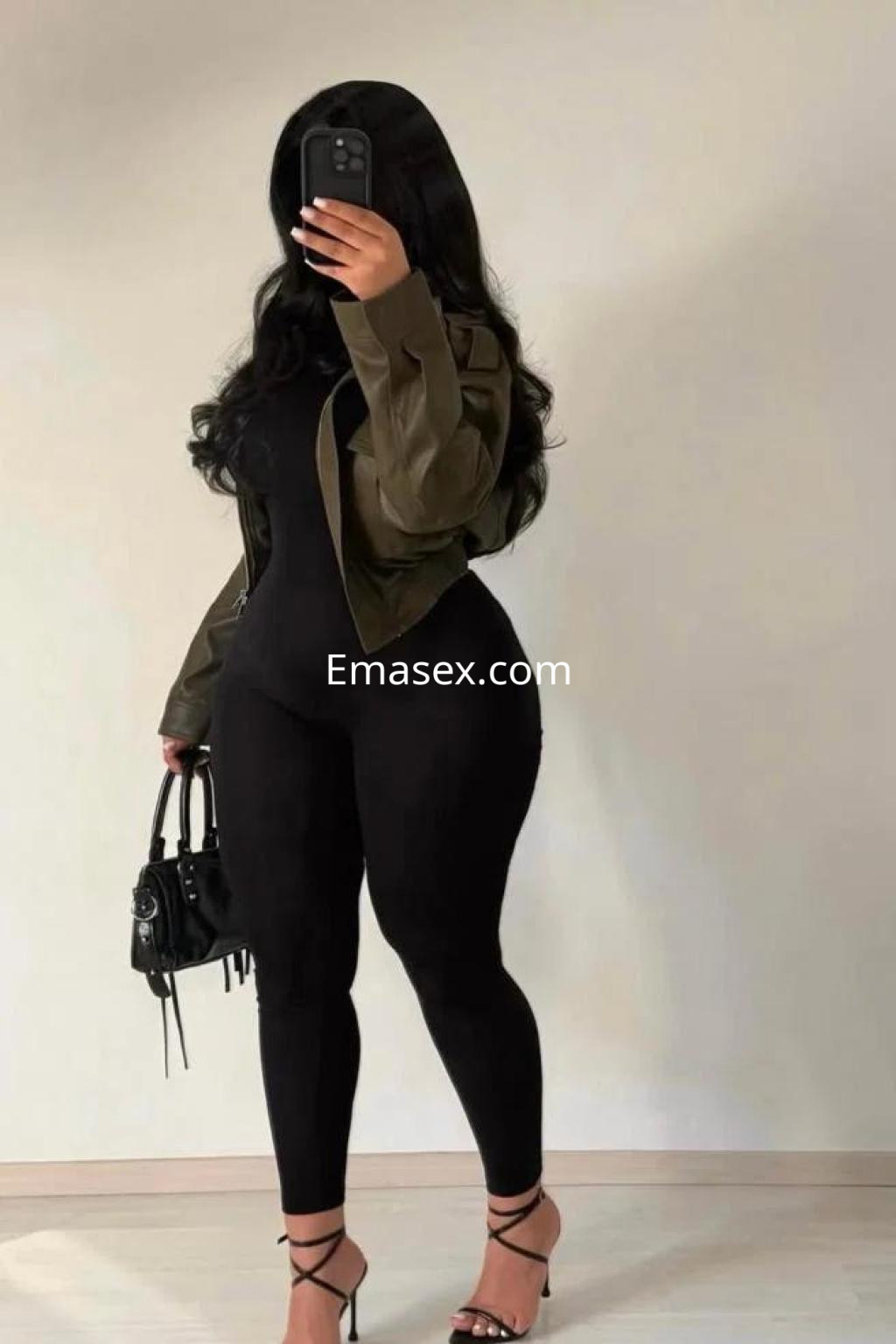 La puta en Murcia viste un ajustado mono negro y tacones, sosteniendo un bolso mientras se mira en el espejo.