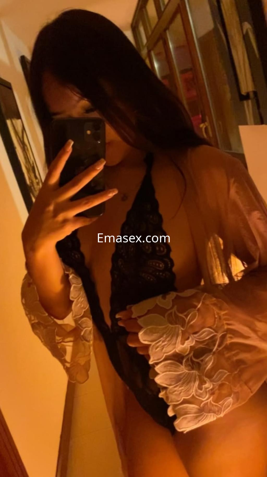 Escort en Donosti, mostrando un body negro y una bata con detalles florales, posando con un espejo en un ambiente tenue.