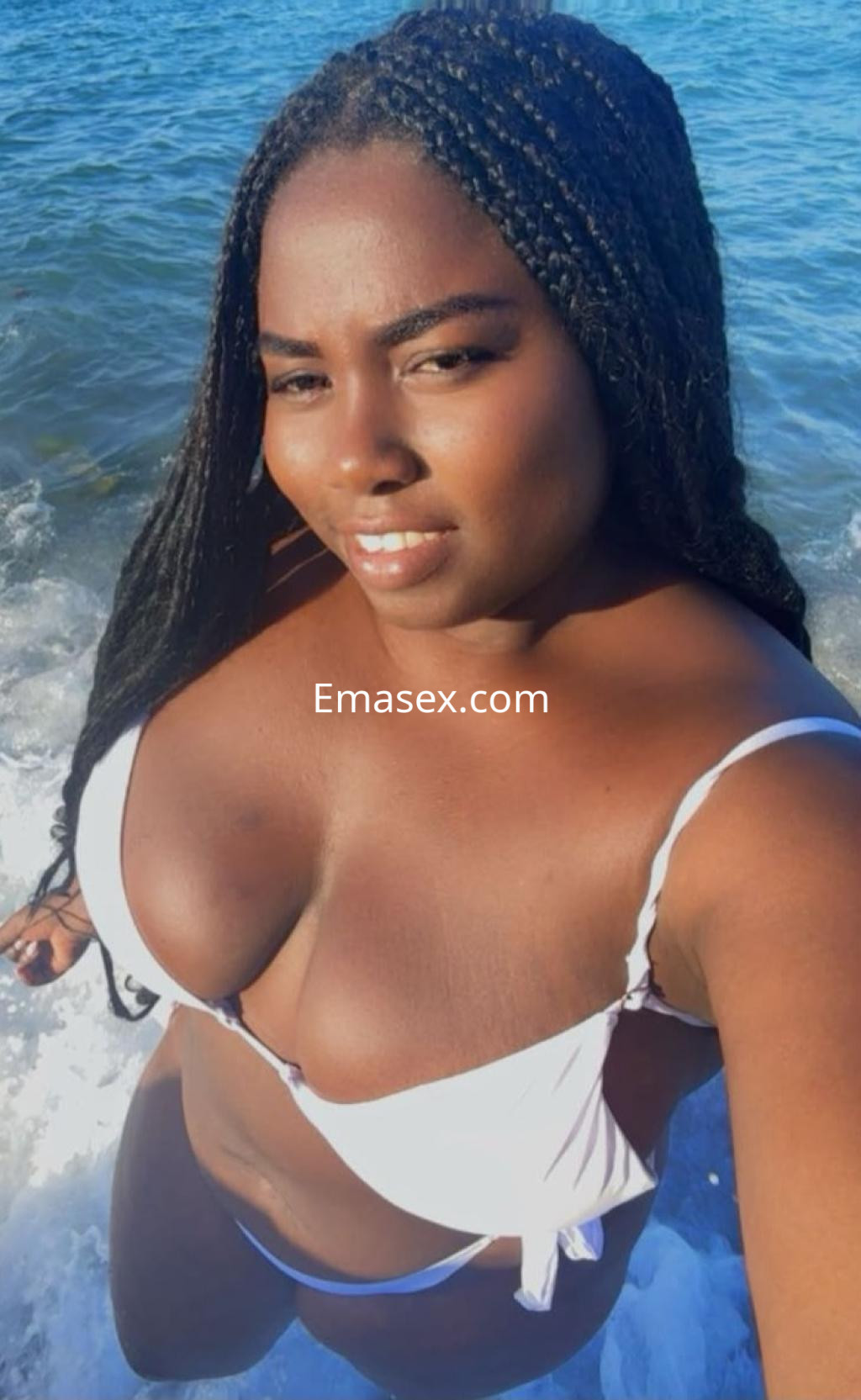 Escort en Granada con piel oscura y cabello trenzado, posando en el agua con un bikini blanco y sonriendo.