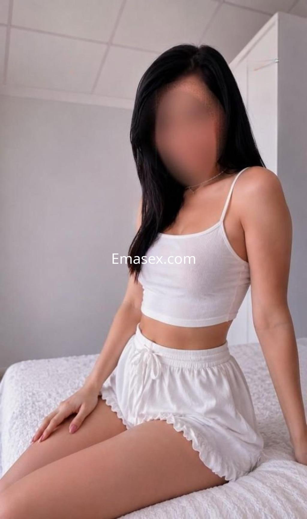 Puta posando en Albacete con blusa blanca sin mangas y pantalones cortos claros, sentada sobre una cama.