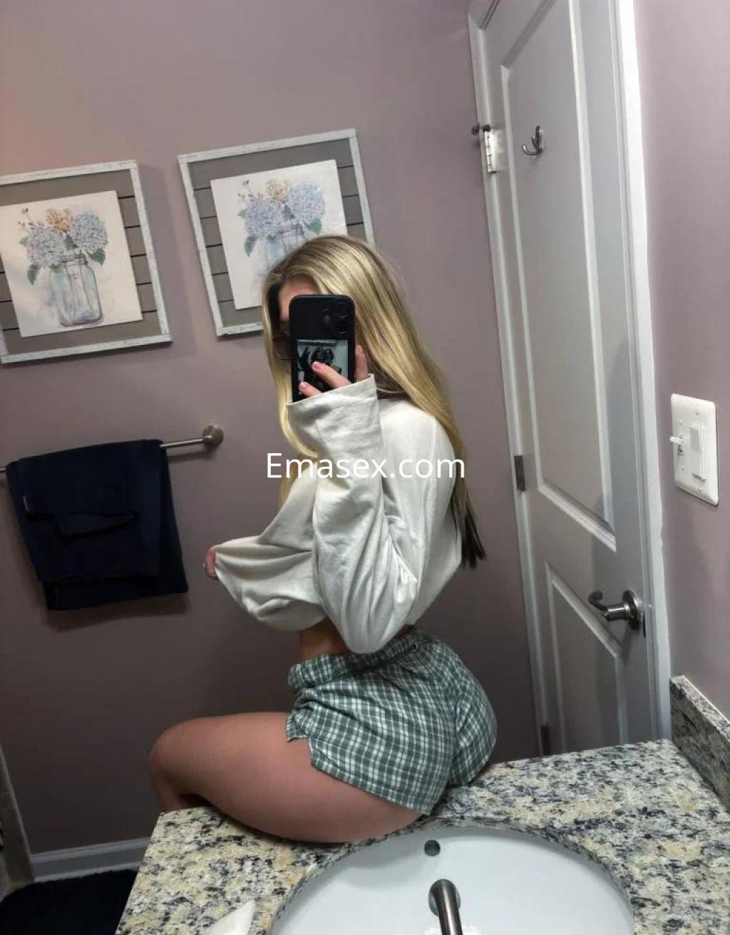 Puta en Cáceres con cabello rubio y largo, usando una blusa blanca y pantalones cortos de cuadros en un baño.