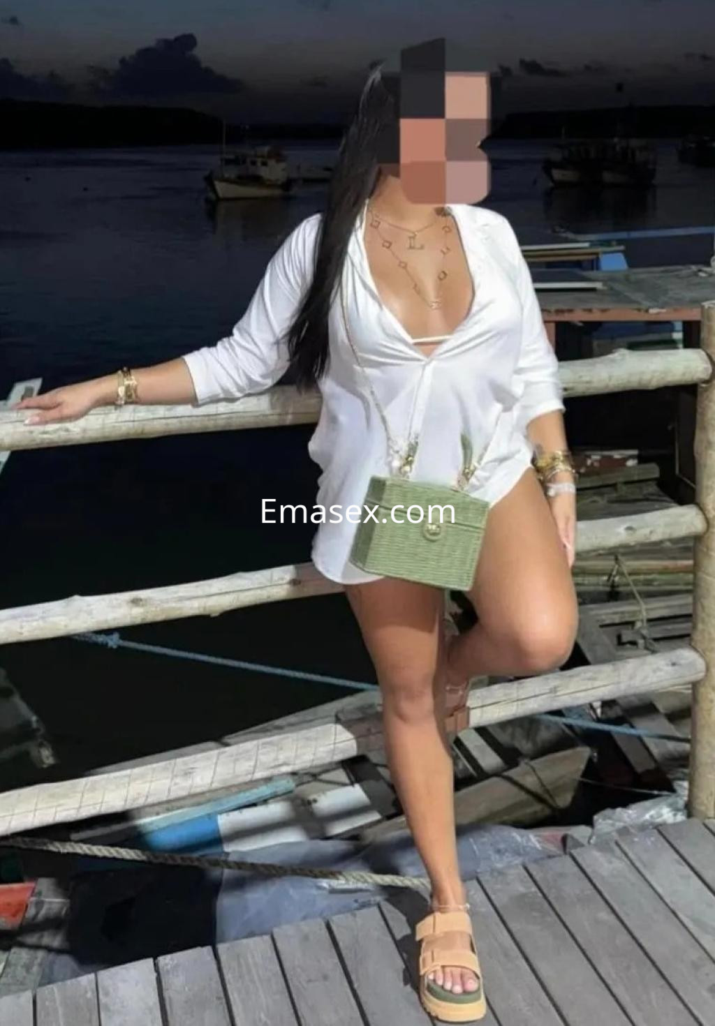 Escort posando en un muelle al atardecer, lleva una blusa blanca y sandalias, con un bolso verde en Oviedo.