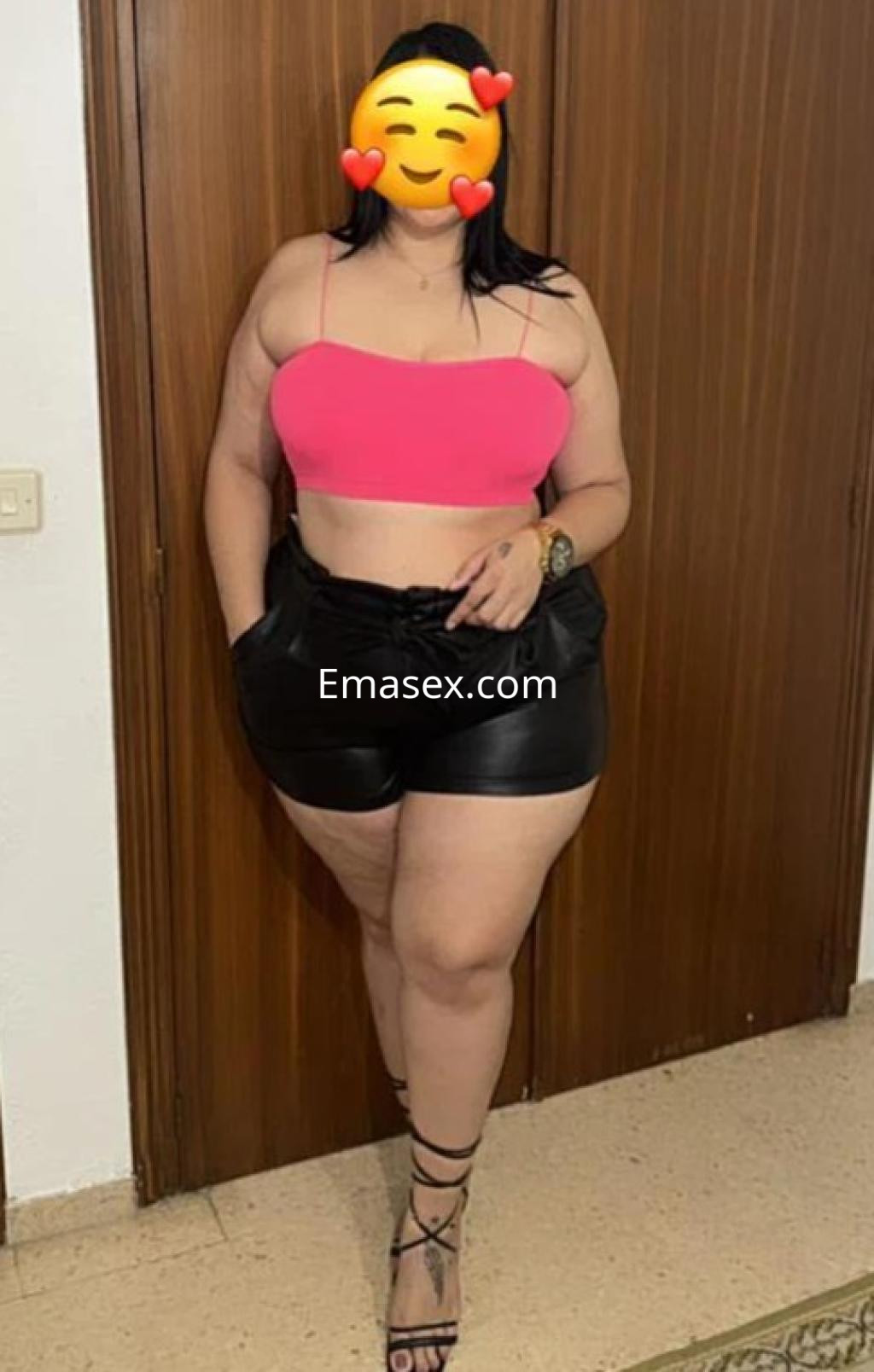 Escort con top rosa y pantalones cortos de cuero, posando con una mano en la cadera en un entorno interior de Valladolid.