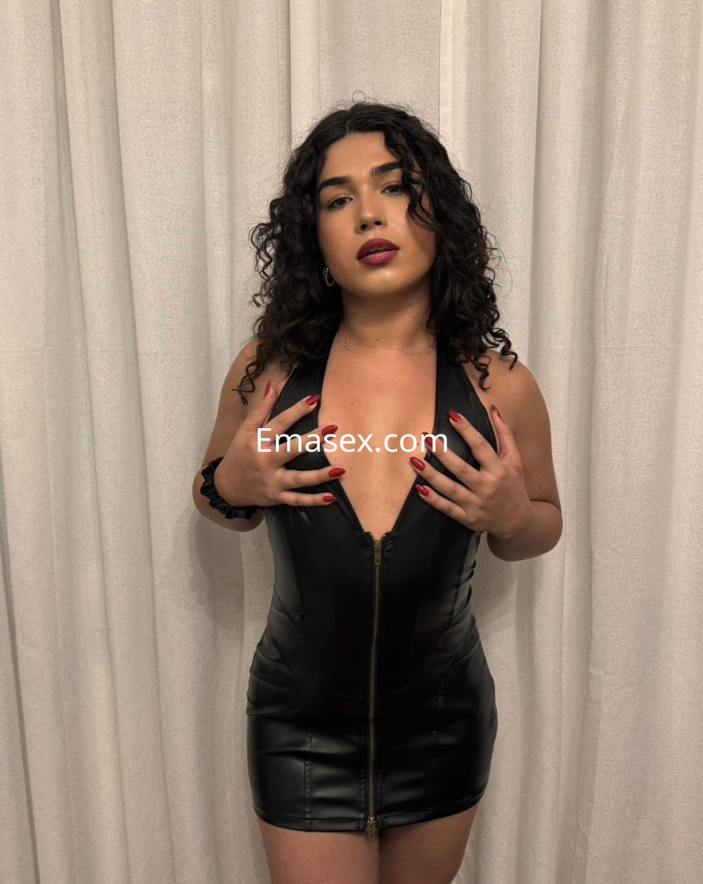 Puta trans en Santander, luciendo un vestido negro ajustado con escote y cabello rizado, posando en un fondo claro.