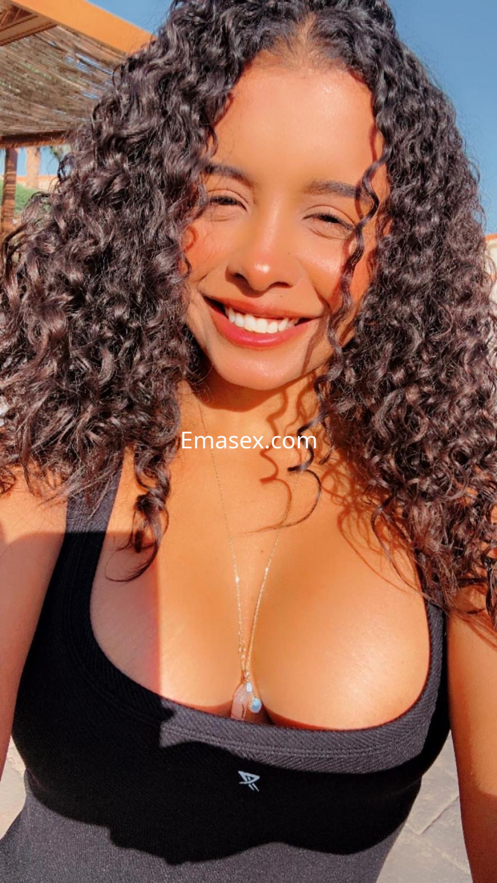 Puta sonriente con cabello rizado y suelto, vistiendo un top negro, en un ambiente soleado de Santander.