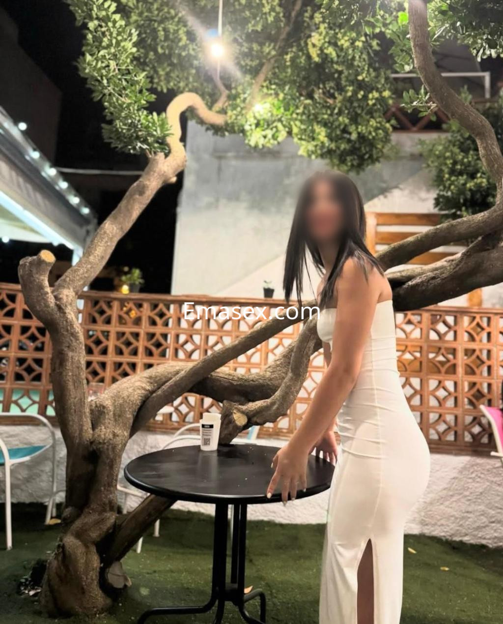 La escort se encuentra en un entorno al aire libre en Pamplona, luciendo un vestido blanco ajustado y posando junto a una mesa.