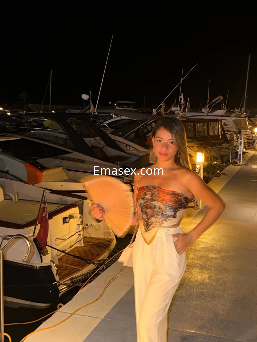 Escort en Palma de Mallorca, con vestido sin mangas y pantalones claros, sostiene un abanico frente a un puerto.