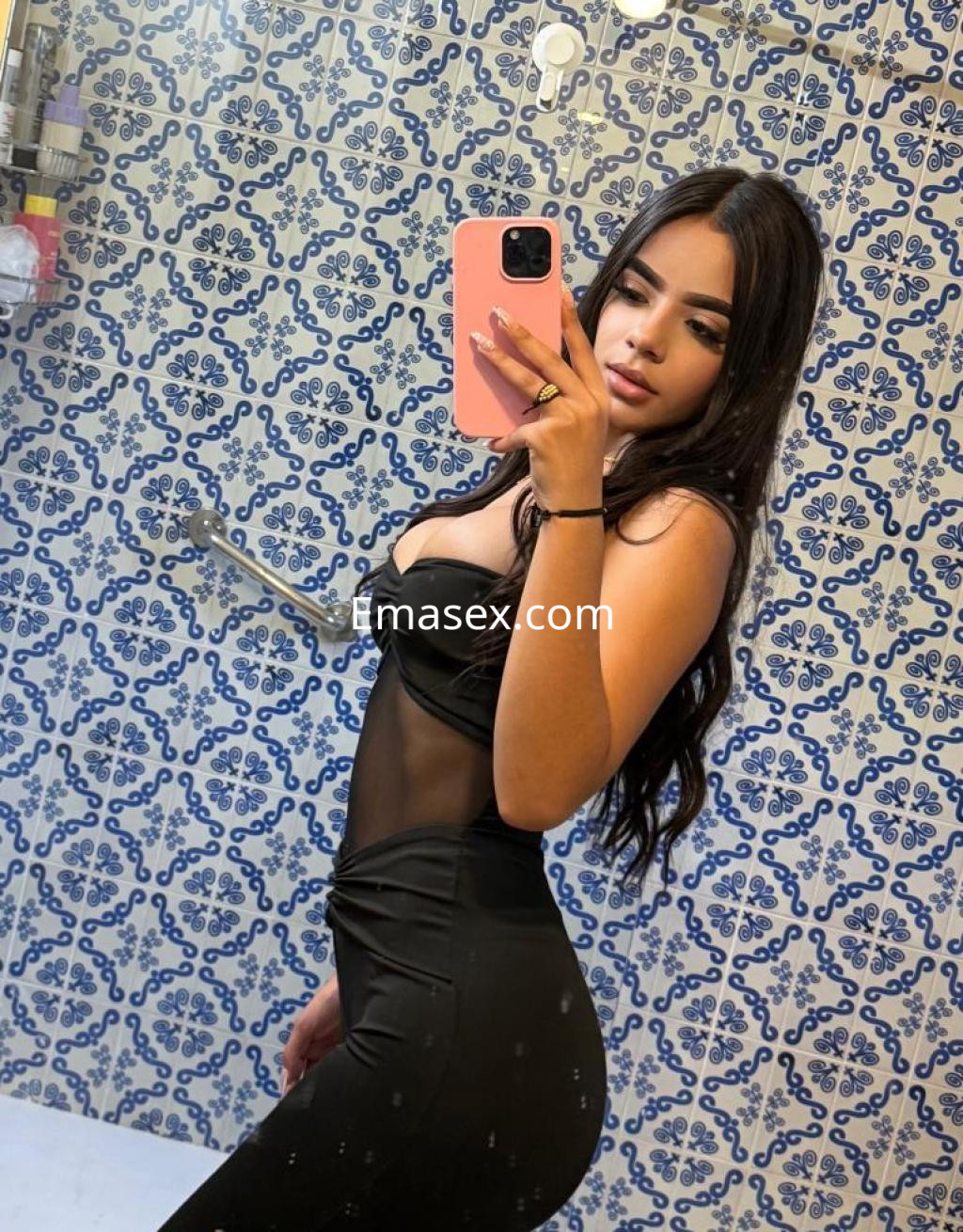 La escort se fotografía en un baño con azulejos azules, luciendo un conjunto negro ajustado y cabello largo.