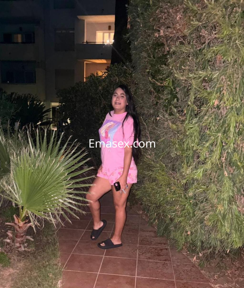 Puta en León posando en un jardín, vestida con camiseta rosa y shorts, luciendo relajada y sonriente.