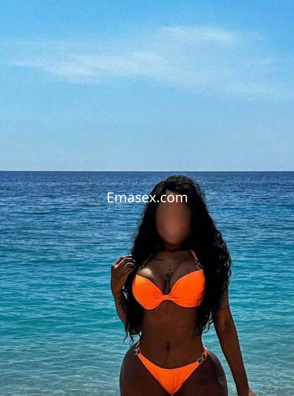 Puta en Granada luciendo un bikini naranja en la playa, con el mar azul y cielo despejado de fondo.