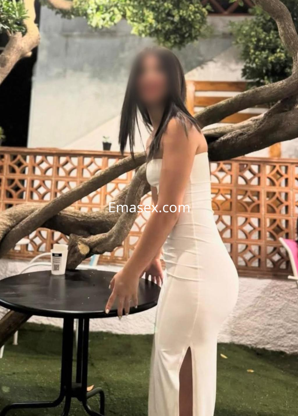 La puta en Pamplona lleva un vestido blanco ajustado con una abertura lateral, posando junto a una mesa.